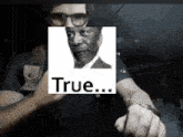a man wearing glasses holds a picture of a man with the word true above it
