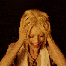 a woman with blonde hair and a necklace holds her hands to her head