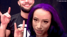 a woman with purple hair is standing next to a man with a beard and giving a peace sign .