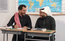 two men sit at desks in front of a map with arabic writing on it