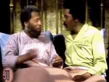 two men are sitting on a bed and talking to each other . one of them is wearing a yellow shirt .