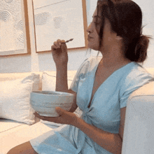 a woman in a blue dress is sitting on a couch eating from a bowl with a spoon
