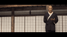 a bald man in a black robe stands in front of a white window
