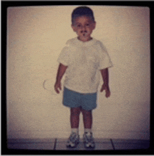 a young boy wearing a white shirt and blue shorts stands in front of a wall