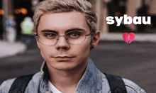 a young man wearing glasses and a denim jacket with the word sybau behind him
