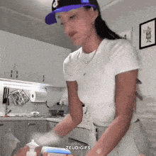 a woman wearing a purple hat and a white shirt is cleaning in a kitchen with the words " zeudigifs " below her