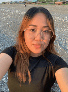 a woman wearing glasses and a black shirt takes a selfie on a rocky beach