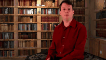 a man in a red shirt sits in front of a bookshelf full of books