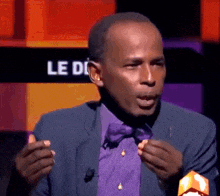 a man in a blue suit and purple bow tie is speaking in front of a sign that says le di