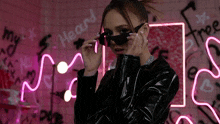 a woman wearing sunglasses is standing in front of a neon sign that says my heart