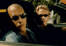 two men are sitting in a car wearing sunglasses and looking out the window .