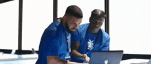 two men in blue shirts are looking at a laptop .