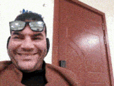 a man wearing sunglasses and ear muffs smiles in front of a red door