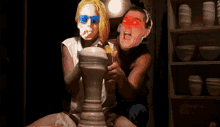 a man and a woman are sitting on a pottery wheel and the woman is wearing blue sunglasses