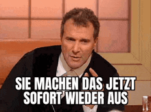 a man is sitting at a table with a sign that says `` sie machen das jetzt sofort wieder aus ''