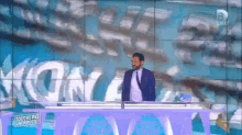 a man in a suit and tie is standing at a desk in front of a sign that says kon
