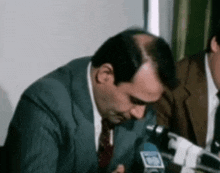 a man in a suit and tie is talking into a microphone while sitting at a table .