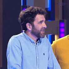 a man with a beard is wearing a blue shirt with a black embroidered logo on the left sleeve