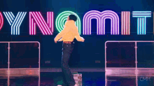 a man in a yellow shirt is dancing on a stage in front of a neon sign that says dynamite .