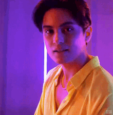 a man in a yellow shirt is standing in front of a purple wall and looking at the camera .