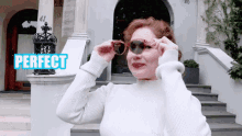 a woman in a white sweater adjusts her sunglasses in front of a sign that says perfect
