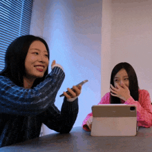 two women are sitting at a table with one holding a cell phone and the other holding a tablet