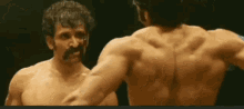 a shirtless man with a mustache looks at another shirtless man in a mirror