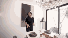 a woman in a black dress is standing in a dining room with a painting on the wall .