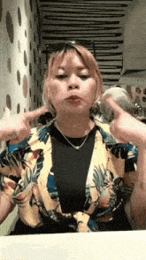 a woman is sitting at a table making a funny face while wearing a floral shirt and sunglasses .