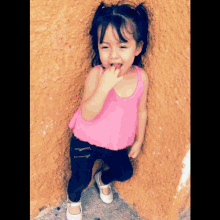 a little girl in a pink tank top and black pants stands next to a yellow wall