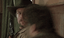 a man with a beard wearing a cowboy hat holds a gun