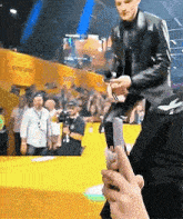 a man in a leather jacket is standing on a stage holding a microphone while a person holds a cell phone .