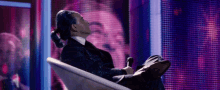 a man in a suit sits in a chair with his legs crossed while holding a microphone