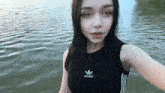 a young woman in a black adidas tank top is taking a selfie in front of a body of water .