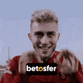 a man in a red shirt is smiling and making a gesture with his hands in front of a betosfer logo .