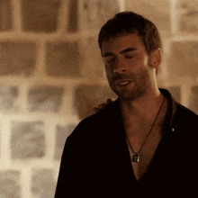 a man wearing a black shirt and a necklace stands in front of a stone wall .