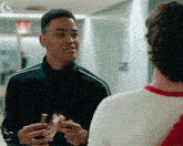 a man in a black jacket is talking to another man in a hallway while holding a candy bar .