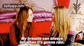 two women are sitting next to each other on a bed and one of them is talking about her breasts .
