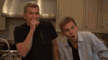 two men are standing next to each other in a kitchen . one of the men is eating something .