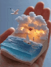 a hand is holding a model of a beach scene with a sailboat in the ocean