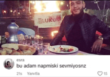 a man sits at a table with a plate of food in front of a sign that says ' kluku '