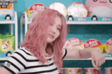 a woman with pink hair and a striped shirt is standing in front of a shelf of ramen cups .