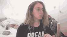 a woman wearing glasses and a black shirt with the word break on it is standing in front of a bed .