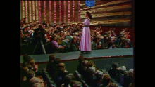 a woman in a pink dress stands on a stage in front of a crowd of people