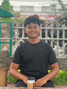 a young man in a black nike shirt sits on a wooden bench holding a cup of coffee