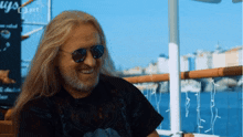 a man with long blonde hair and a beard wearing sunglasses and a black shirt with the word guys on it