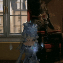 a woman in a blue dress is standing in front of a window while a man sits in a chair