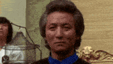 a man with gray hair and a blue shirt is looking at the camera in front of a bird cage .