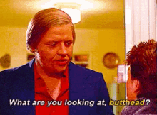 a man in a suit is talking to another man in a red shirt who is asking him what he is looking at