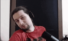 a man wearing headphones and a red shirt is sitting in front of a microphone and looking at the camera .